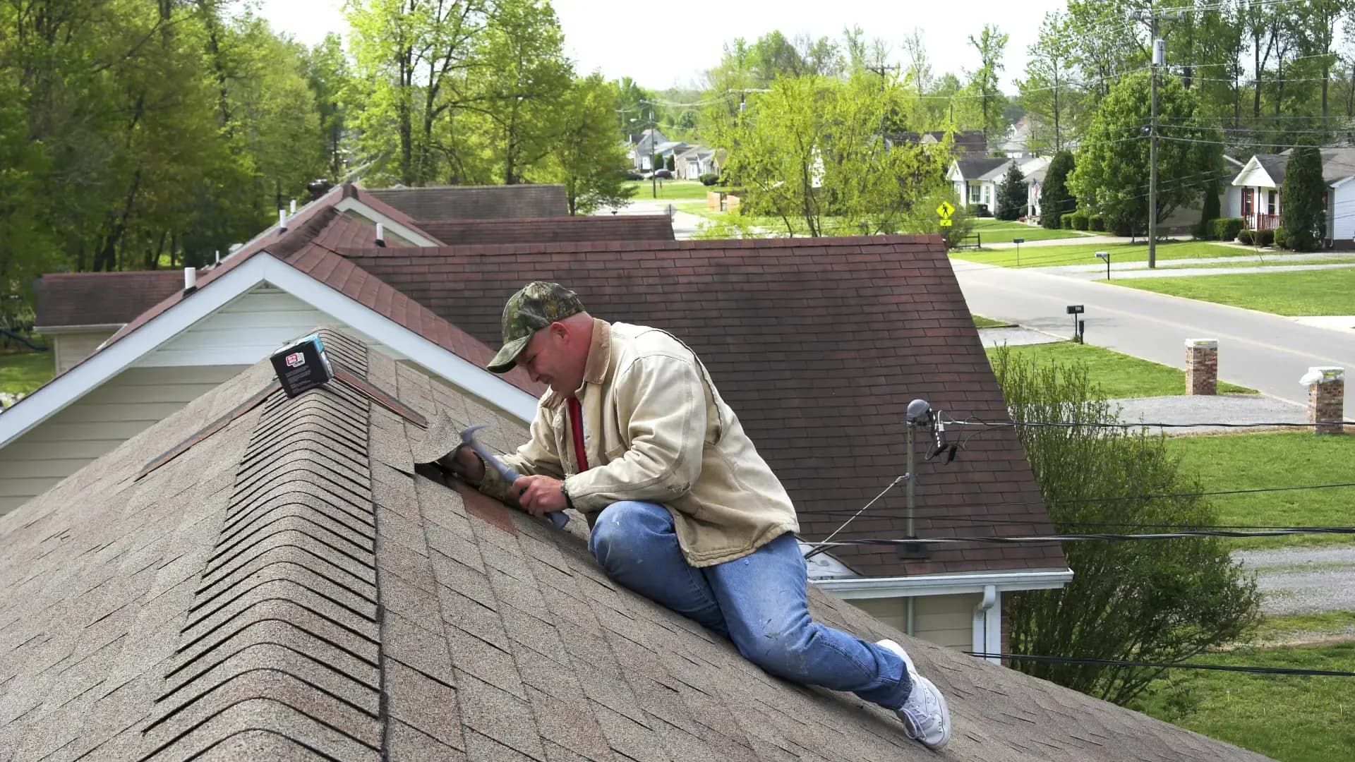 A roofer wearing a camouflage cap, beige jacket, and blue jeans crouches on a residential shingle roof, using a hammer to repair or install ridge cap shingles along the peak, overlooking a quiet suburban neighborhood.