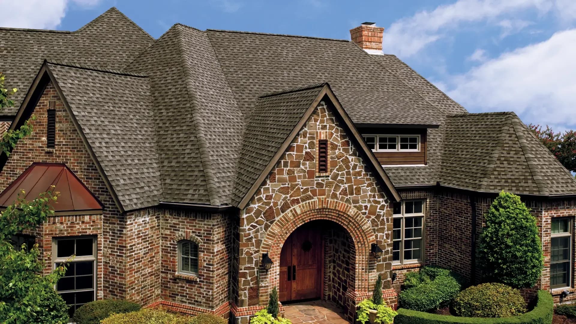 A large, elegant brick house with a complex, gabled roof covered in grey shingles. The entrance features an arched stone facade, framed by manicured bushes.
