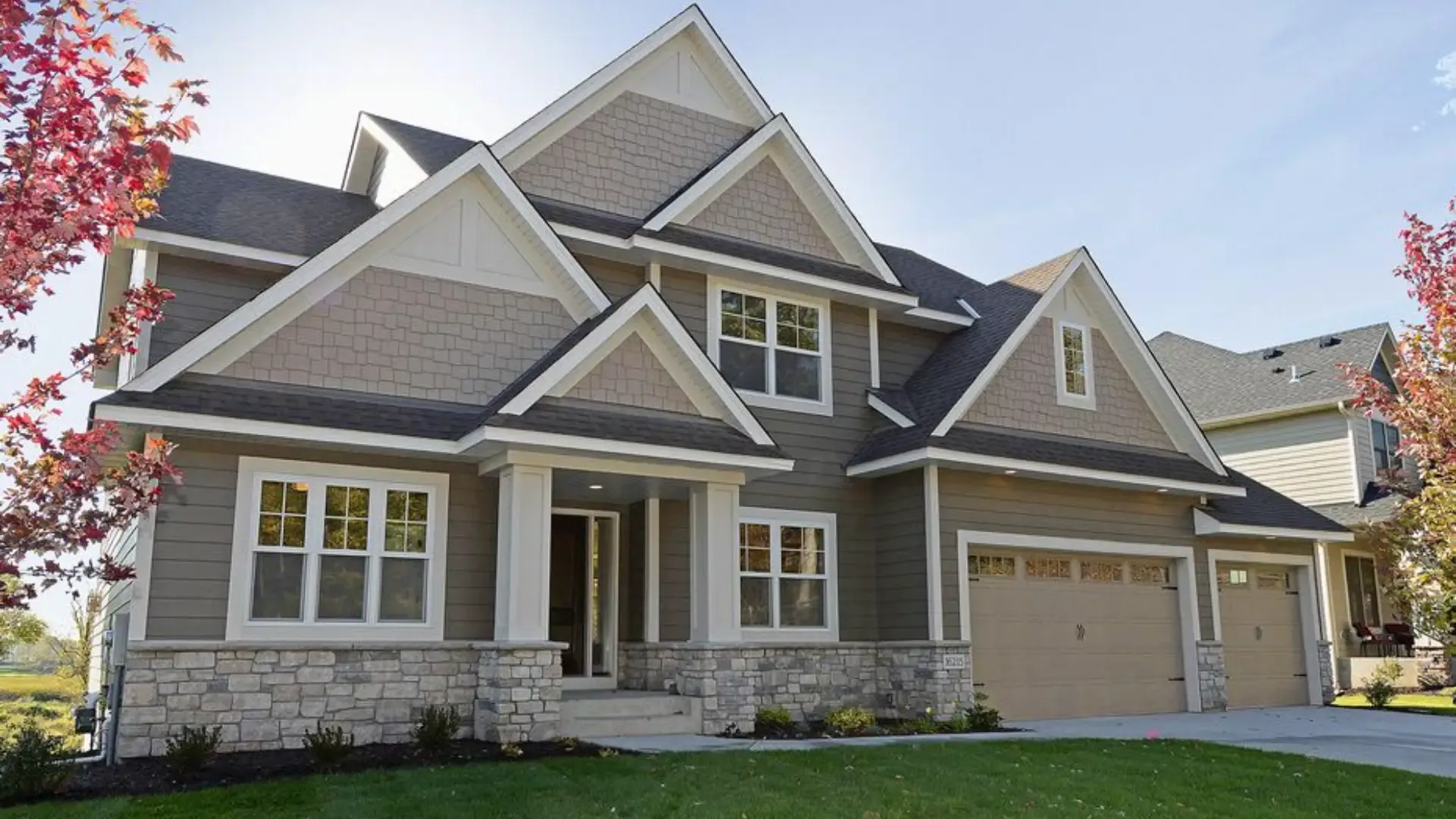 A modern suburban two-story house with gray siding and stone accents. It features multiple gables, large windows, a double garage, and a neat lawn, surrounded by autumn trees.