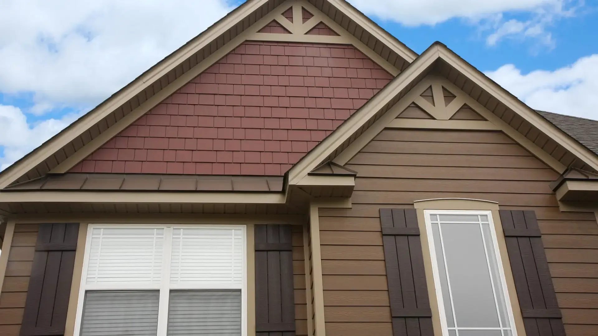 Upward perspective of a Craftsman-style home exterior displaying a combination of reddish-brown shake siding in the upper gable peak and brown lap siding below, highlighted by decorative gable trusses and dark brown board-and-batten shutters.
