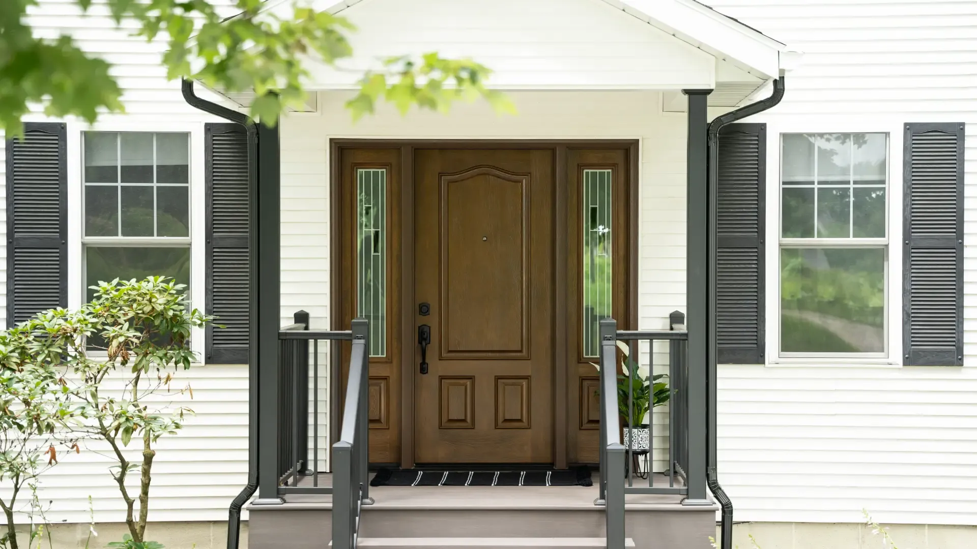 Close-up of a house entrance with a wooden door, flanked by two windows with black shutters. A small porch and green plants add a welcoming feel.