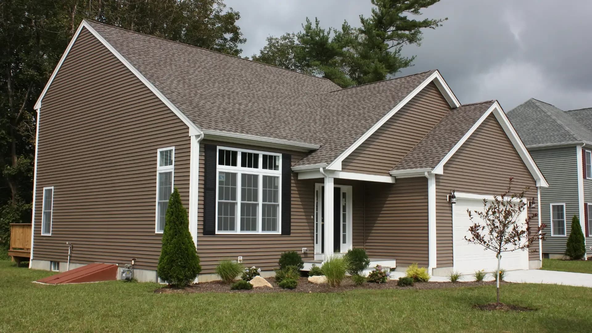 A brown single-story house with a grey roof, large front windows, and a two-car garage. A manicured lawn and small shrubs enhance the peaceful suburban setting.