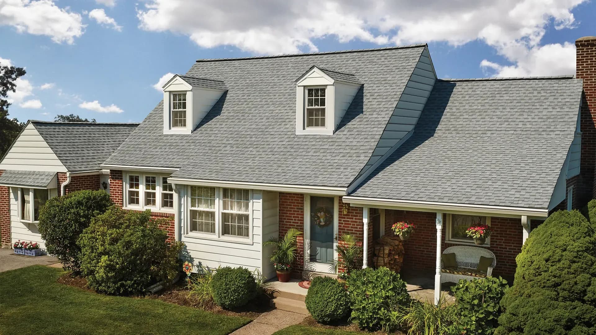 Charming suburban house with a blue-gray gabled roof, white dormer windows, and a brick facade. Lush greenery and bright blue sky enhance its inviting appearance.