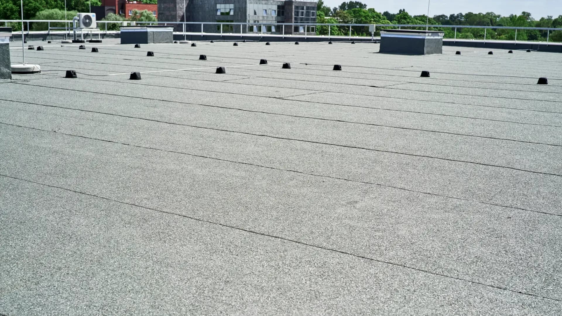Flat rooftop with grey, textured surface, dotted with small black fixtures and air conditioning units. Green trees and buildings are visible in the background.