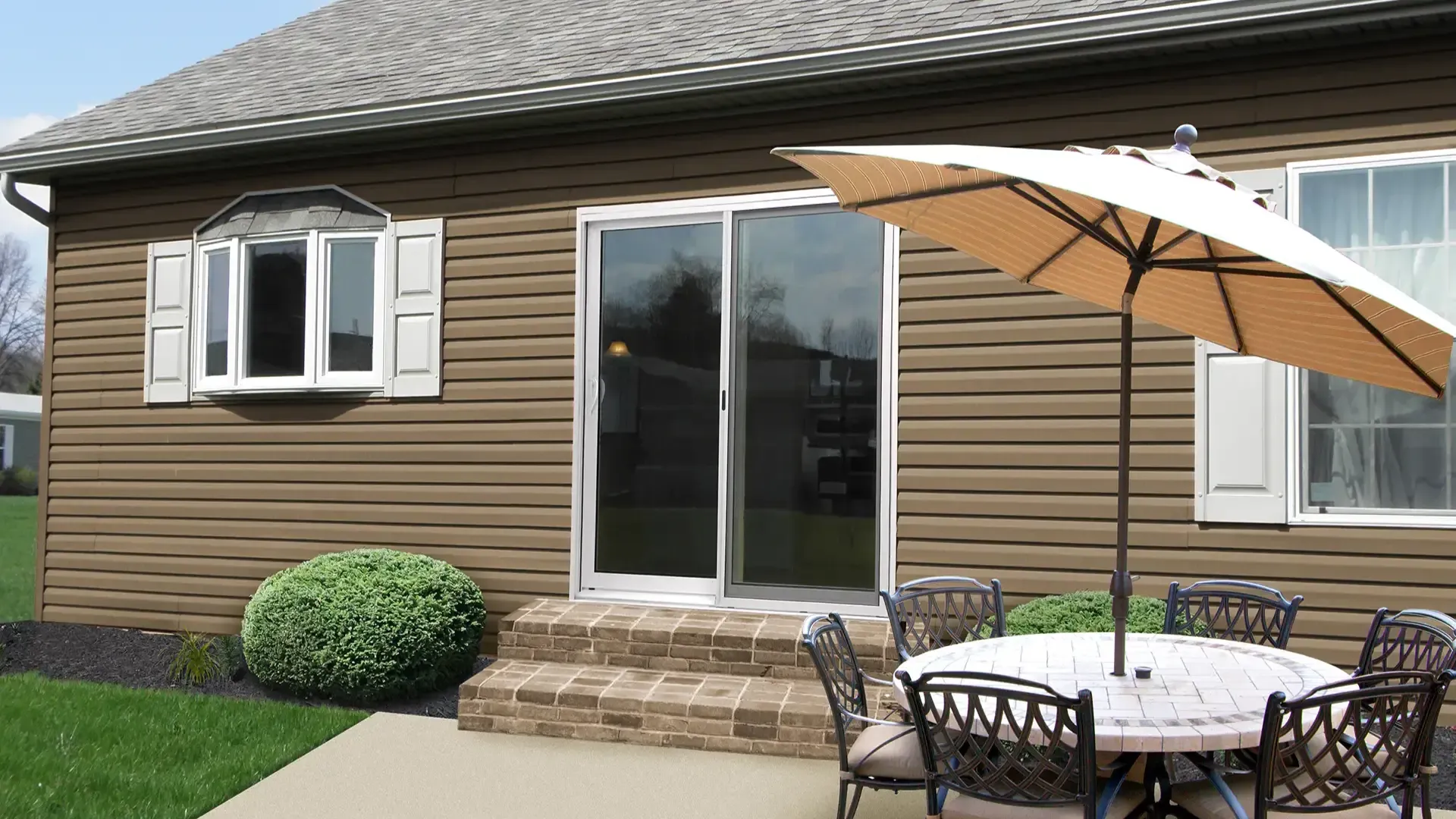 A patio with a round table and chairs under a large umbrella sits on a concrete surface. The backdrop is a brown house with white-framed windows.