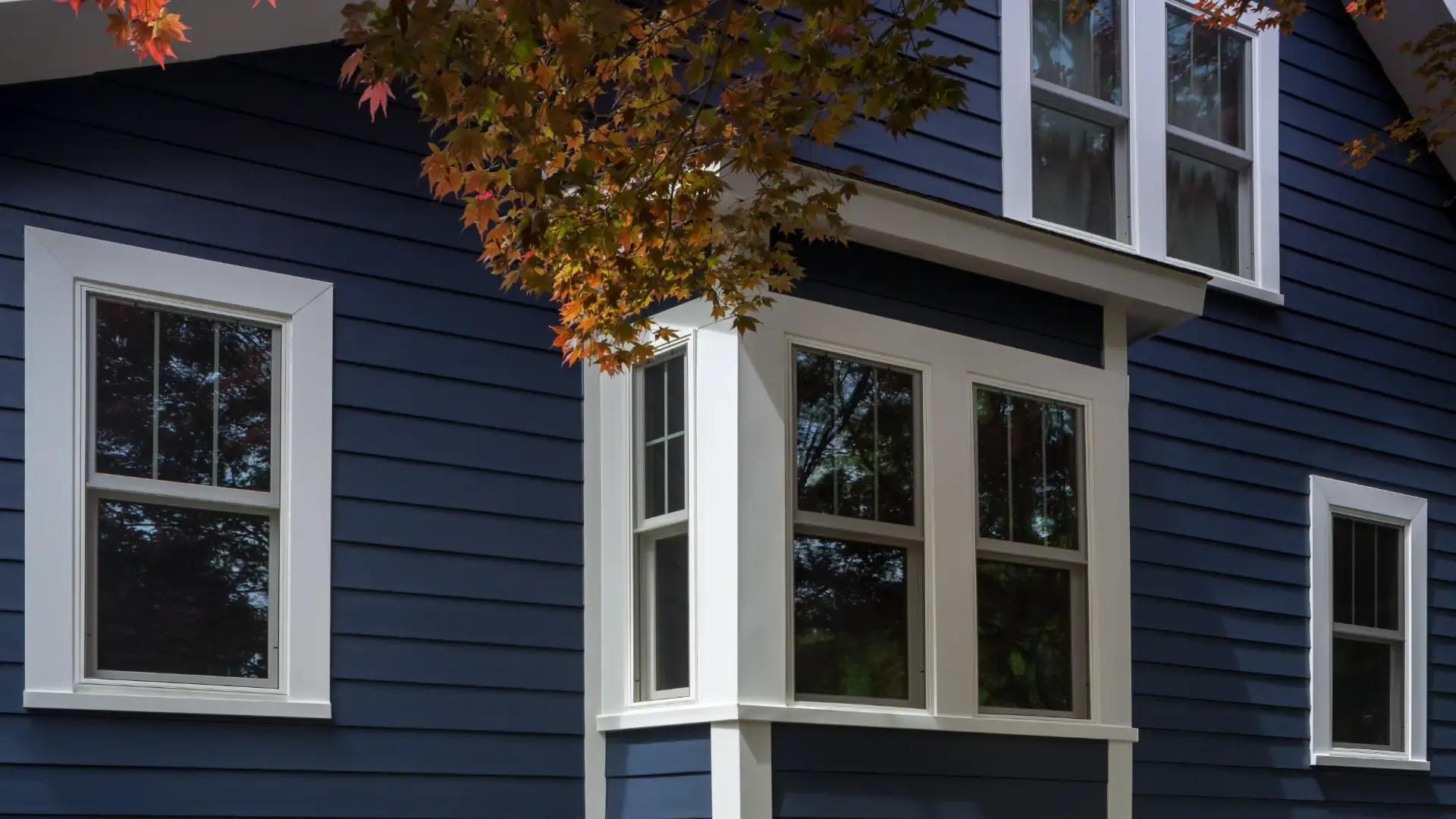 A deep blue house with crisp white-trimmed windows stands under the shade of a tree with red leaves, creating a serene and inviting atmosphere.