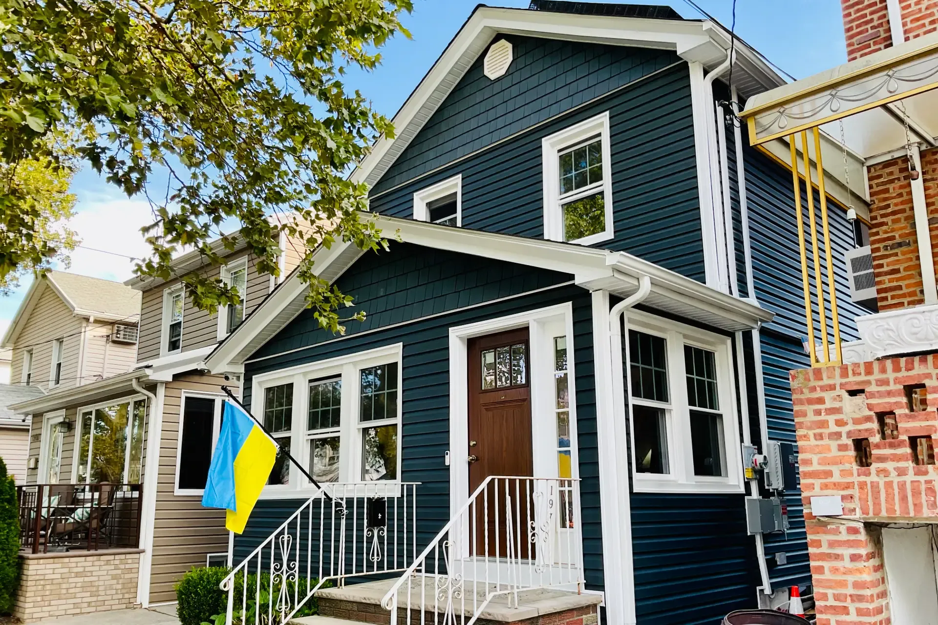 A dark blue house with white trim and a brown door, displaying a Ukrainian flag. The house has several windows and is surrounded by other homes and trees, conveying a peaceful neighborhood vibe.