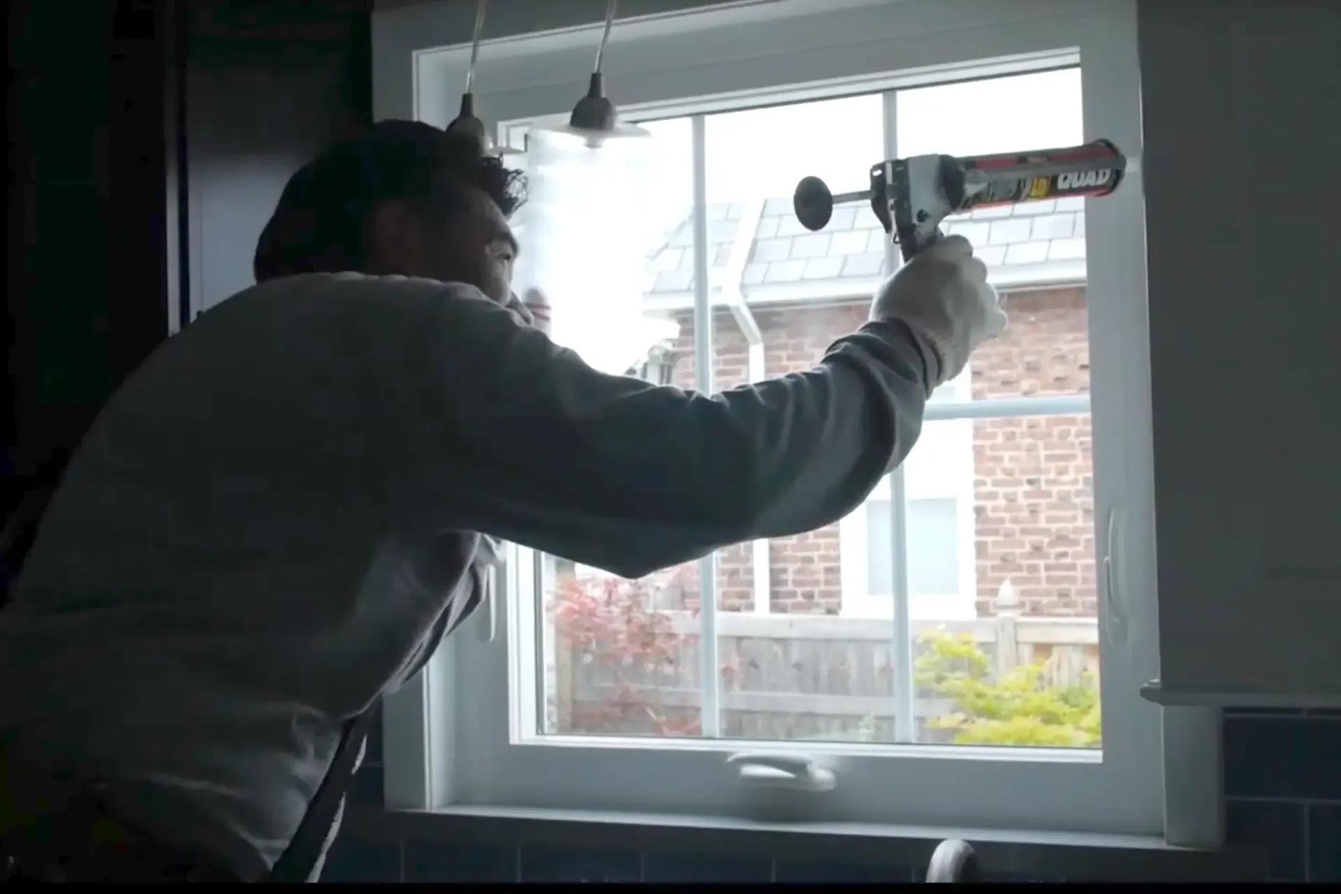 A person is sealing a window with a caulking gun. They're focused, wearing a dark hoodie and gloves. Outside, a brick house and garden are visible.