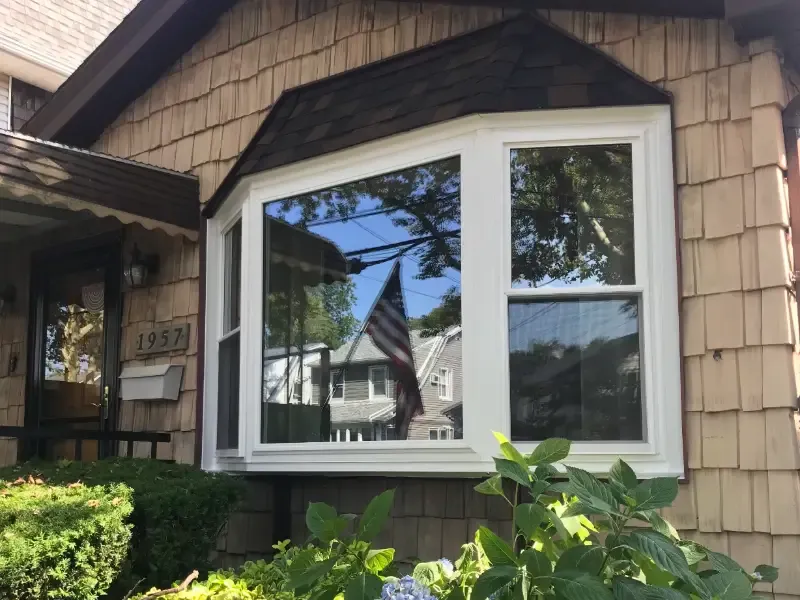 A bay window on a shingle-style house reflects trees and a waving American flag. The facade features number 1957. Lush greenery grows in front.