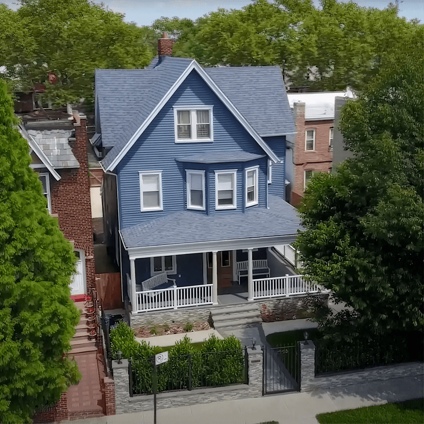 Aerial view of a charming blue house with white trim, a steep roof, and a cozy porch. Surrounded by lush trees, conveying a serene neighborhood feel.