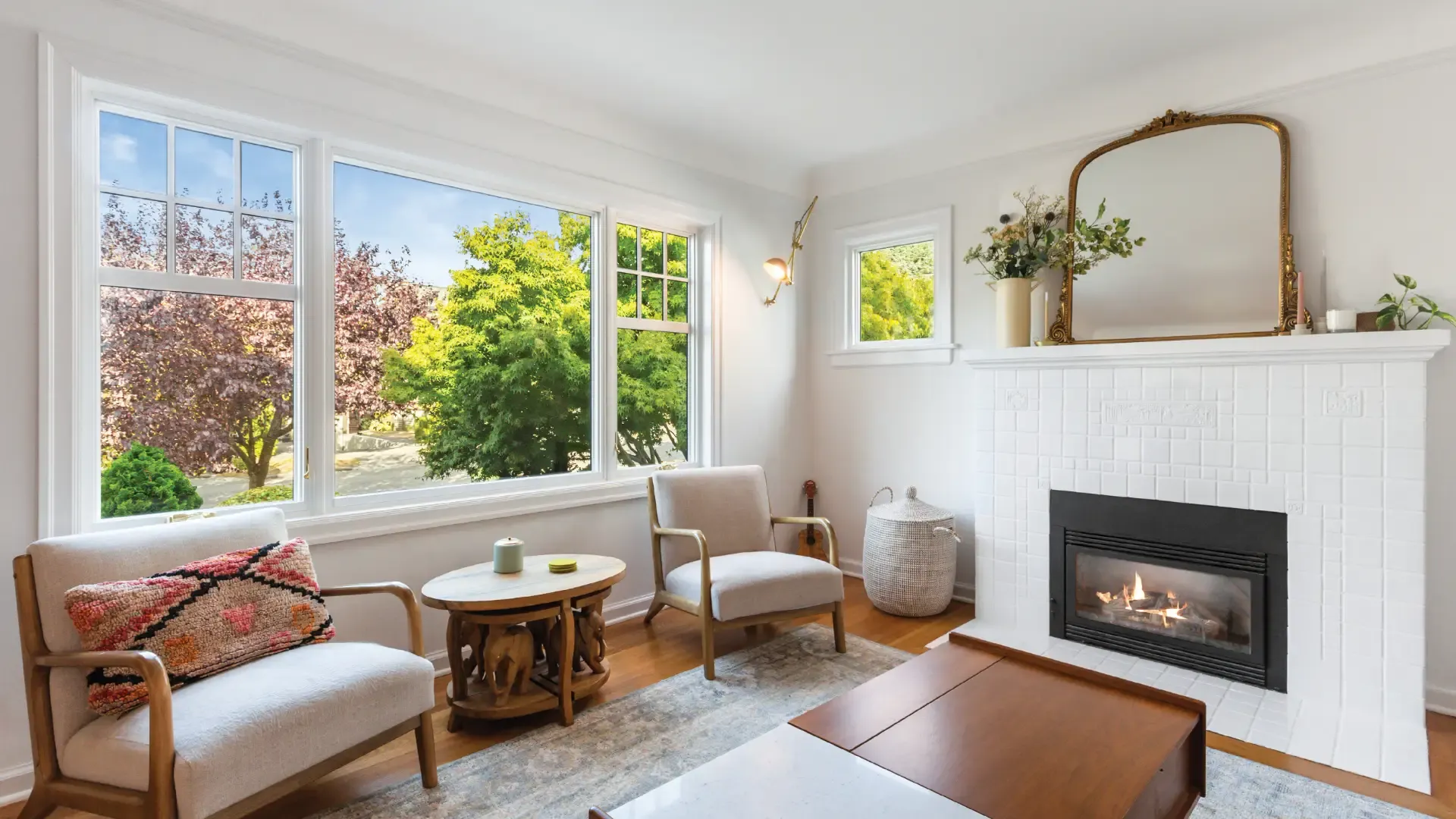 Bright living room with large windows revealing lush green trees outside. Features cozy armchairs, a wooden coffee table, and a lit fireplace.