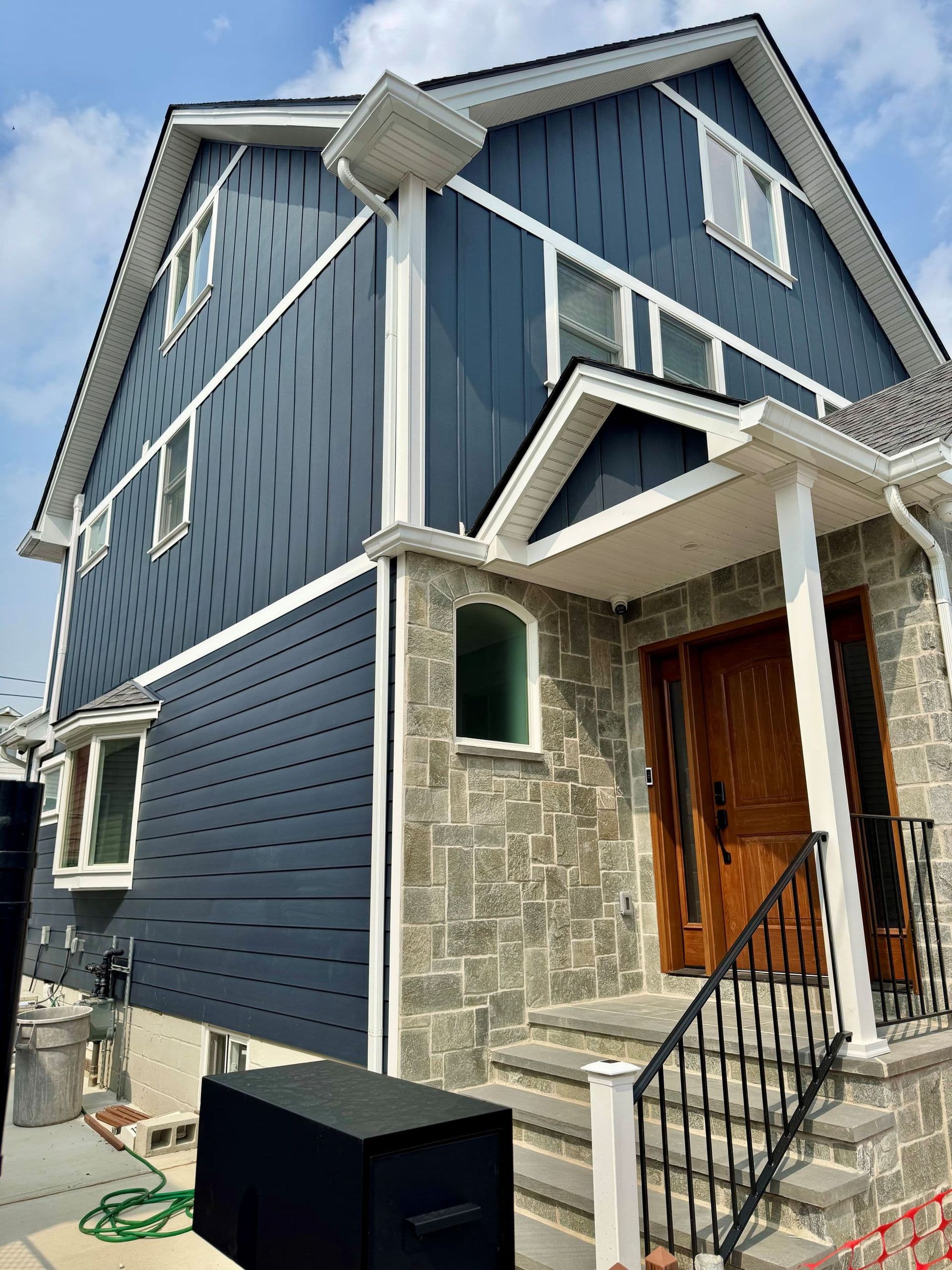 A modern two-story house with blue siding and a stone facade around the entrance. It features a wooden front door, white trim, and a gabled roof under a clear sky.