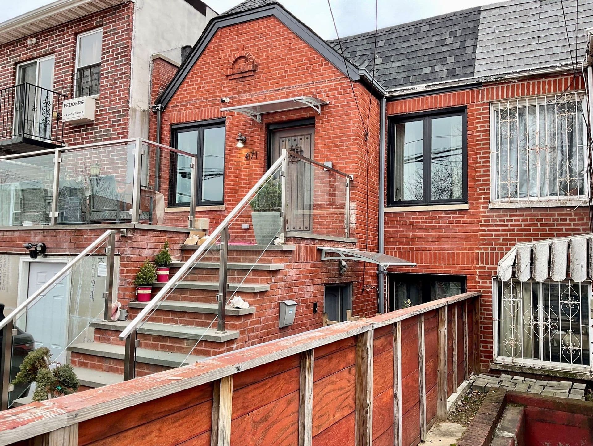 Modern red brick townhouse with a glass-railed staircase. It features large windows, potted plants on the steps, and a cozy front porch.