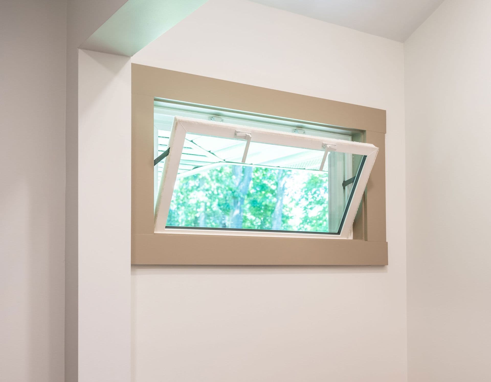 A rectangular awning window partially open, framed in beige, within a white wall. Green trees are visible outside, suggesting a fresh, airy atmosphere.
