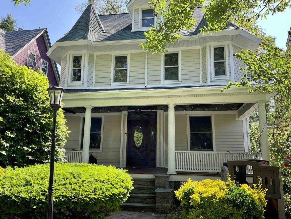 Victorian house with a steep roof, large porch, and dark wooden door, surrounded by lush greenery. Bright, sunny day adds a welcoming feel.