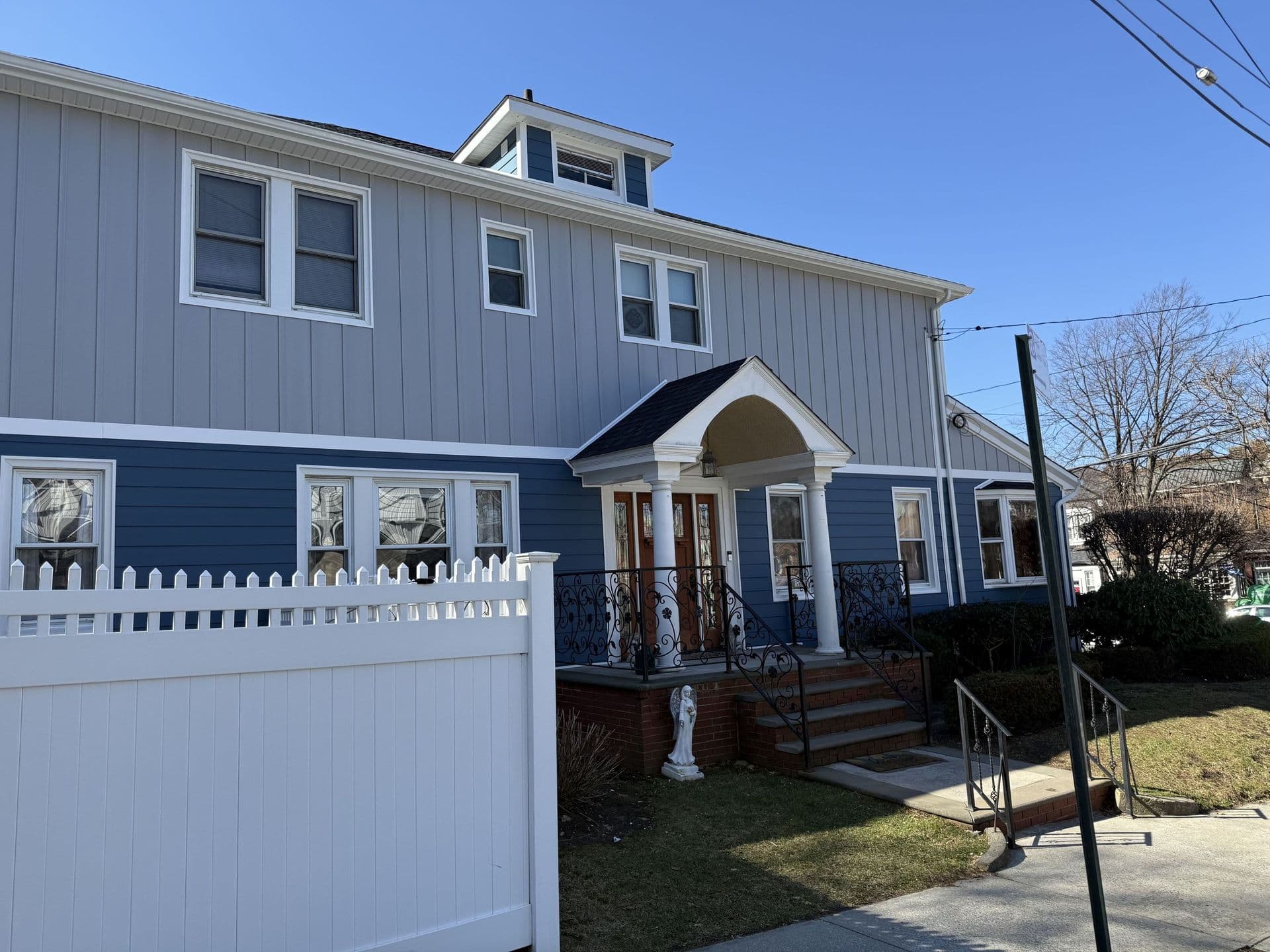 Exterior front view of a two-story residential building featuring light grey vertical siding on the top level and dark blue horizontal lap siding on the main floor, fronted by a white picket fence and a gabled porch entry with white columns.