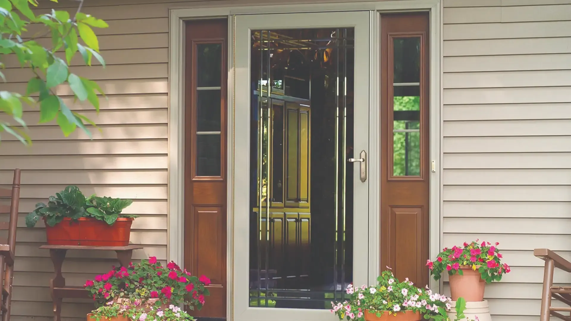 Off-White Storm Door with Decorative Glass and Sidelites (imgi_1_68623b975774fdb0b7cc0c70_decorator2 (1).webp) Exterior view of an entry system set against light beige lap siding, flanked by potted pink and white flowers and wooden rocking chairs. At the center is an off-white storm door featuring full-length glass with decorative brass accent lines and a brushed nickel lever handle. Behind the storm door sits a medium-toned wood main entrance door, flanked by two matching wood sidelights with rectangular upper glass panes.