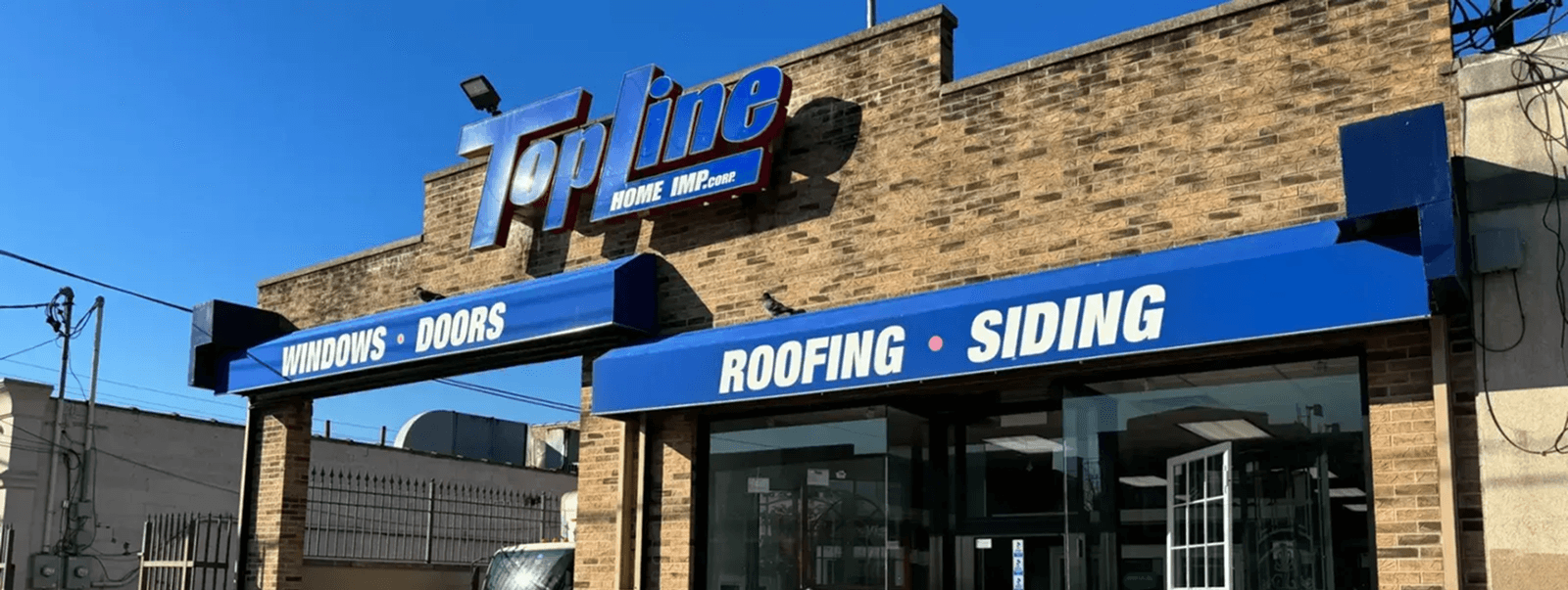 Bright, sunny daytime view of the Topline Home Improvement brick building with blue signage advertising windows, doors, roofing, and siding.