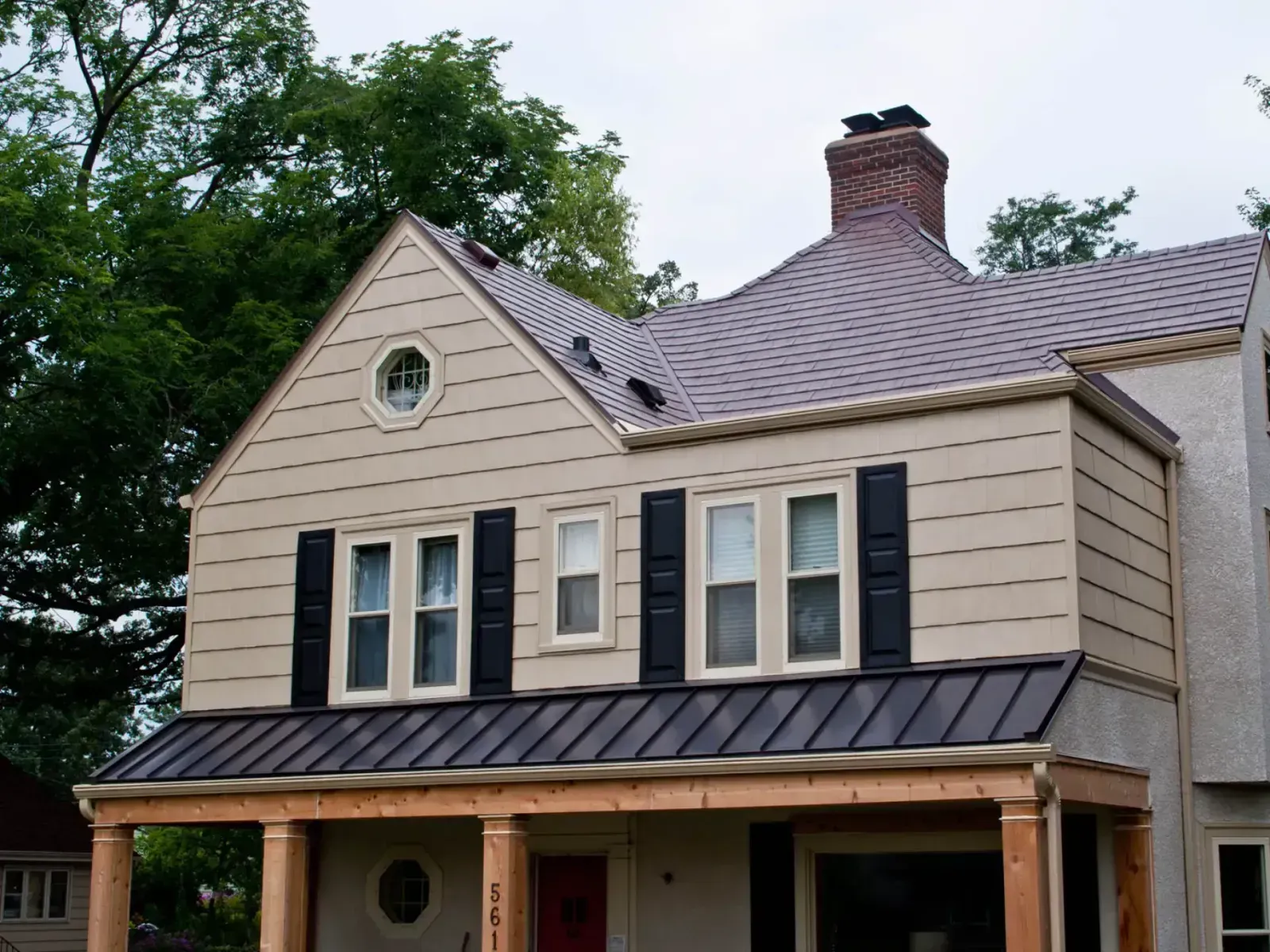 Two-story beige home with black shutters, a dark standing-seam metal porch roof, and a brick chimney.