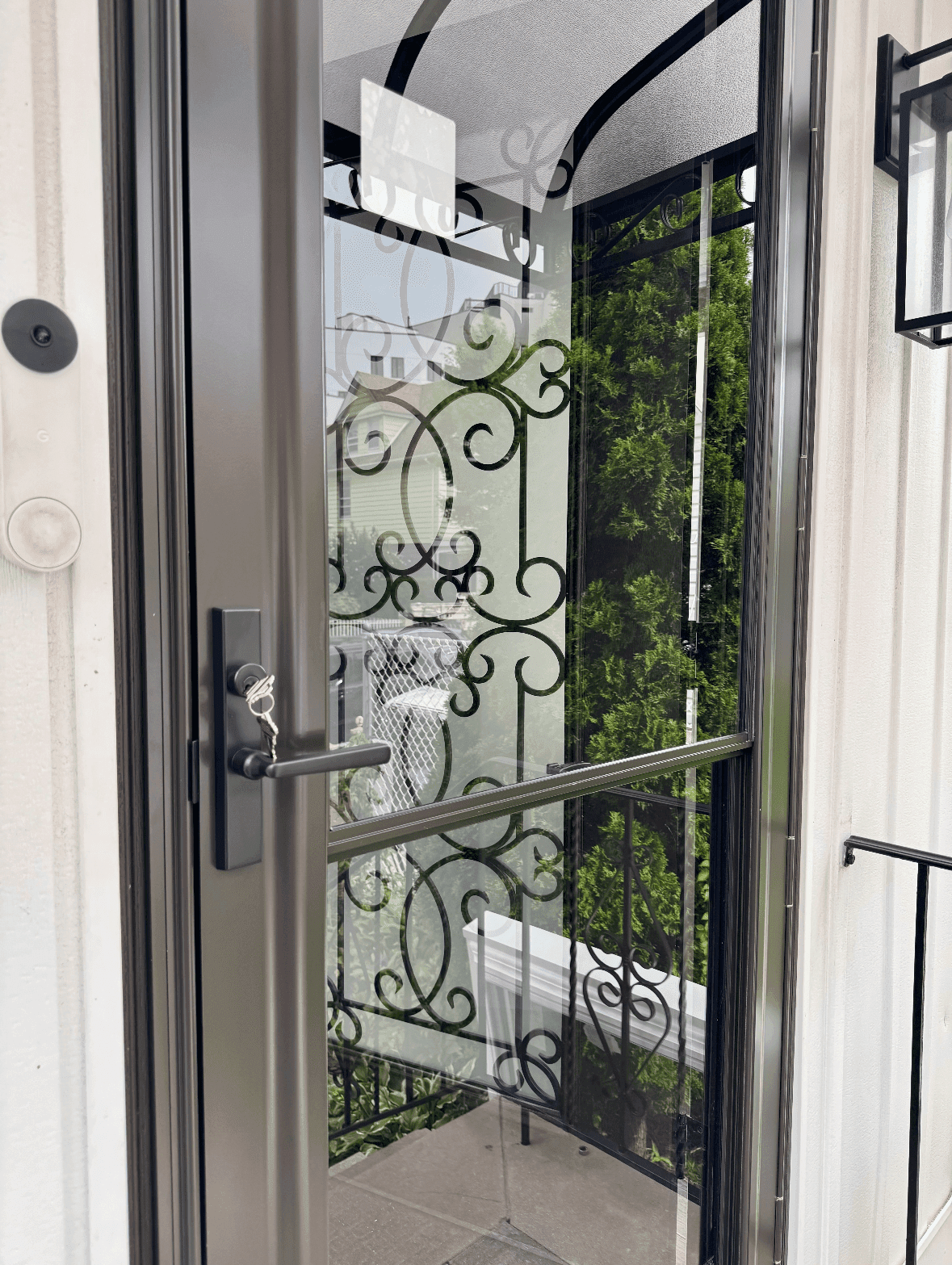 Close-up exterior view of a dark bronze storm door featuring a black lever handle with inserted keys and a full glass panel reflecting decorative wrought-iron scrollwork railings, white siding, and a smart video doorbell mounted on the door trim.