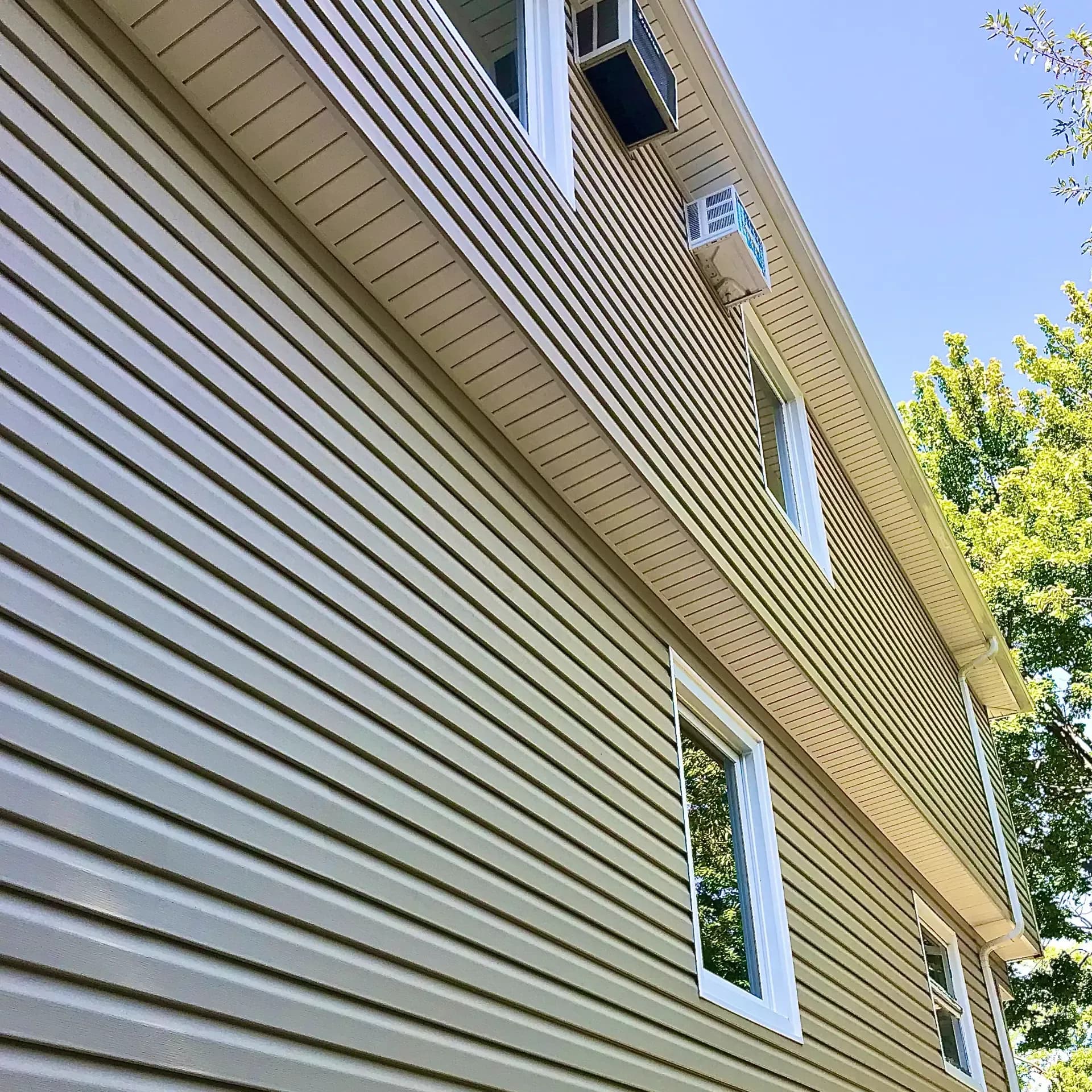 Low-angle exterior shot looking up at the side of a home featuring light beige vinyl lap siding, white window frames, and window air conditioning units against a clear blue sky.